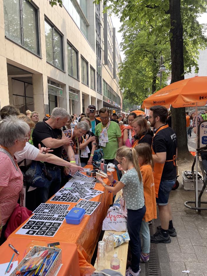 Menschen am orangenen Tisch der Kolpingjugend interessieren sich für die angebotene Aktion 'Airbrush Tattoos'.
