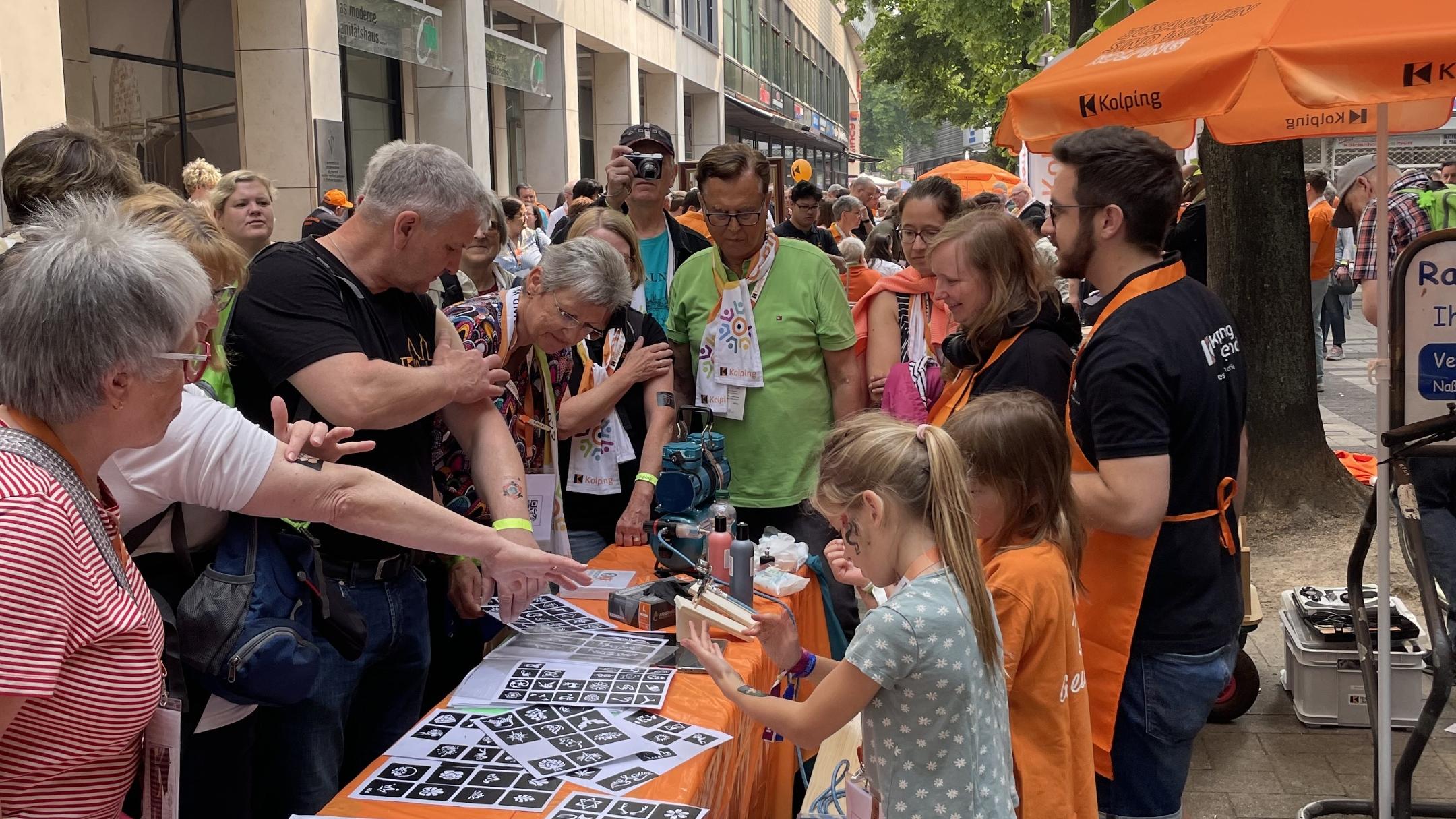 Menschen am orangenen Tisch der Kolpingjugend interessieren sich für die angebotene Aktion 'Airbrush Tattoos'.