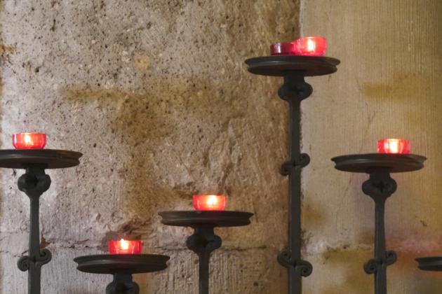 Vor einer Kirchensäule stehen fünf Kerzenständer in unterschiedlicher Höhe mit roten Kerzen.