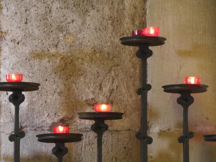 Vor einer Kirchensäule stehen fünf Kerzenständer in unterschiedlicher Höhe mit roten Kerzen.