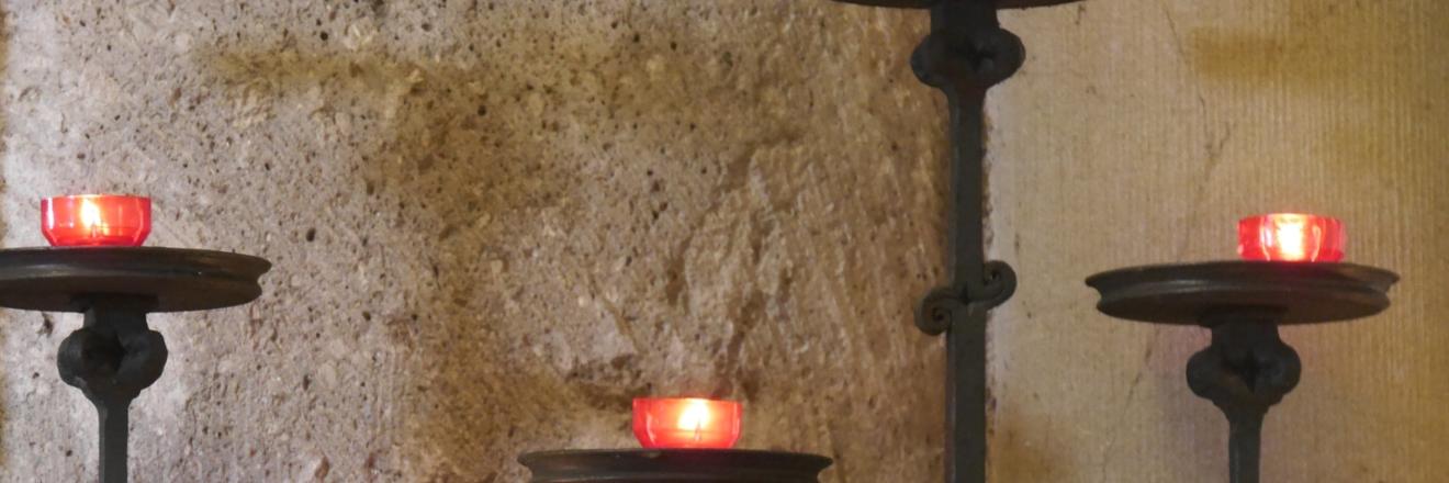 Vor einer Kirchensäule stehen fünf Kerzenständer in unterschiedlicher Höhe mit roten Kerzen.