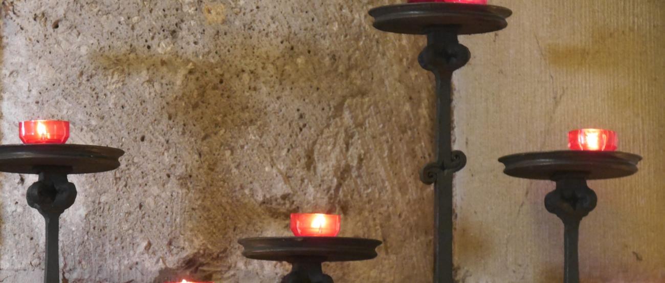 Vor einer Kirchensäule stehen fünf Kerzenständer in unterschiedlicher Höhe mit roten Kerzen.