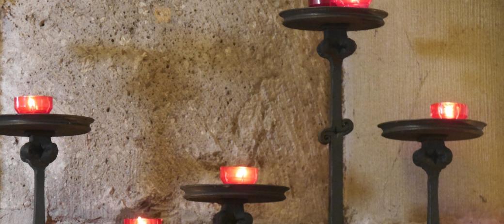 Vor einer Kirchensäule stehen fünf Kerzenständer in unterschiedlicher Höhe mit roten Kerzen.