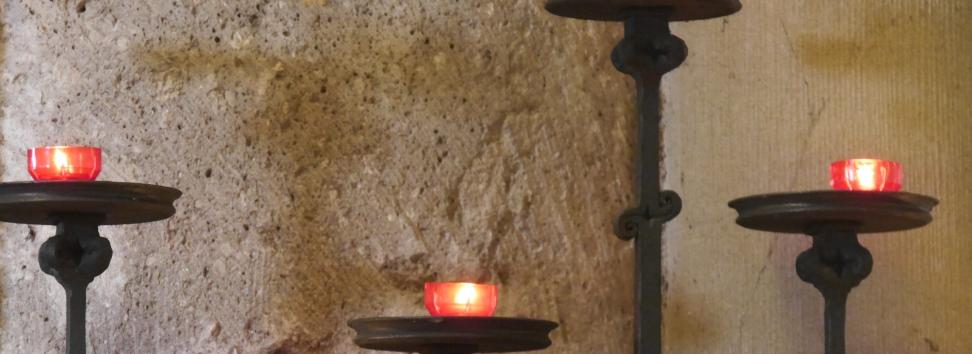 Vor einer Kirchensäule stehen fünf Kerzenständer in unterschiedlicher Höhe mit roten Kerzen.