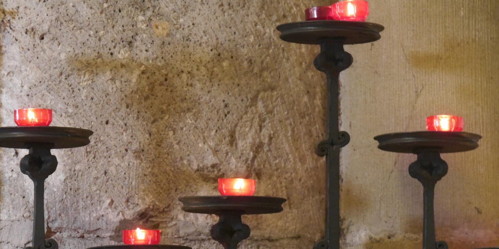 Vor einer Kirchensäule stehen fünf Kerzenständer in unterschiedlicher Höhe mit roten Kerzen. (c) Martina Wagner Vor einer Kirchensäule stehen fünf Kerzenständer in unterschiedlicher Höhe mit roten Kerzen.