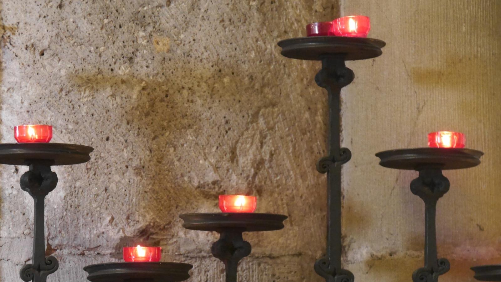 Vor einer Kirchensäule stehen fünf Kerzenständer in unterschiedlicher Höhe mit roten Kerzen.