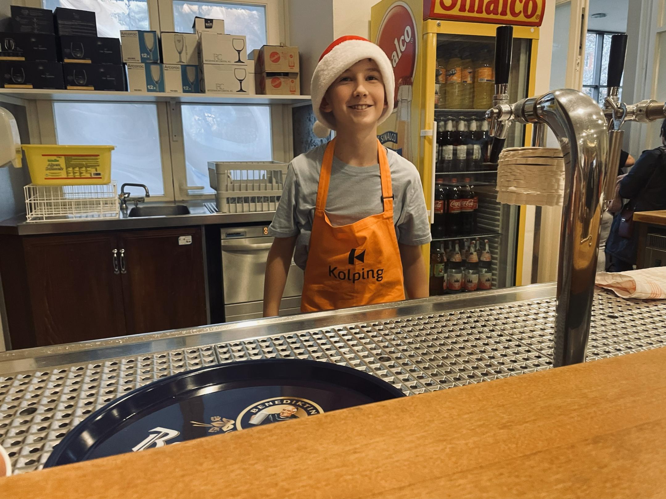 Kleiner Junge mit Kolpingschuerze steht hinter der Theke und hilft bei der Aktion