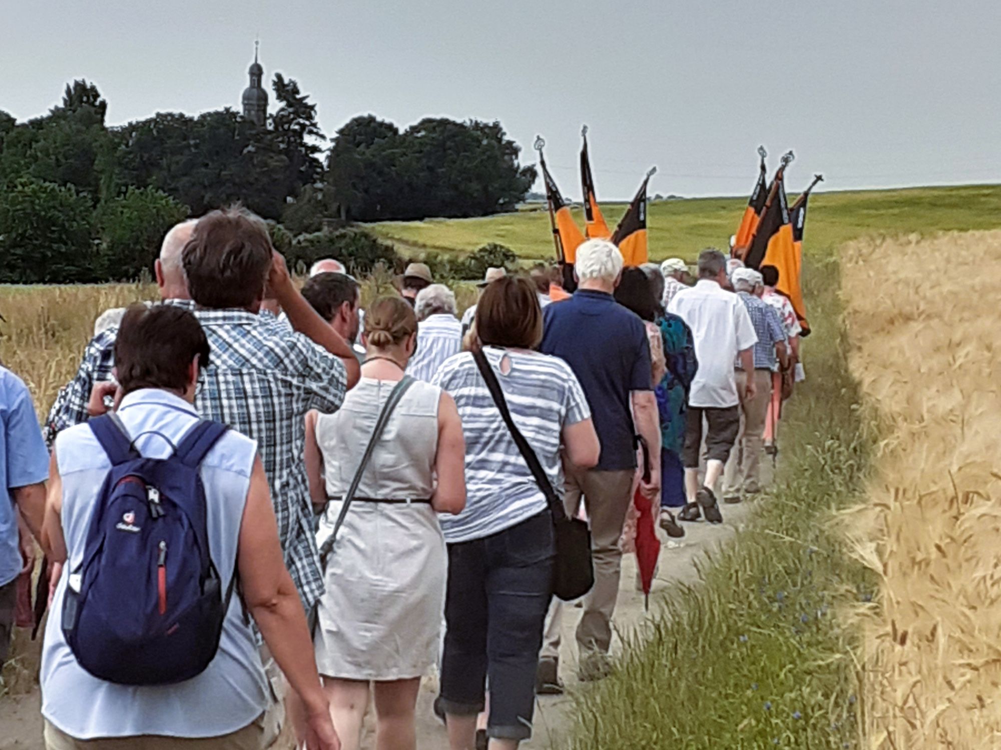 Man sieht eine Gruppe, die mit Kolping-Bannern voran auf einem Feldweg zu einer kleiner Kirche hinter Bäumen pilgert.
