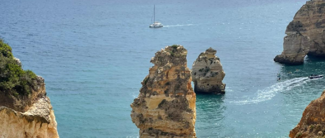 Blick auf das Meer. Man sieht blauen Himmel, Felsen und ein Segelboot.