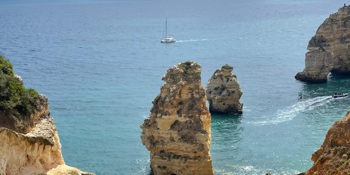 Blick auf das Meer. Man sieht blauen Himmel, Felsen und ein Segelboot.