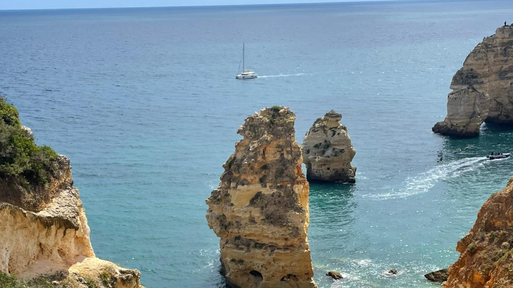 Blick auf das Meer. Man sieht blauen Himmel, Felsen und ein Segelboot.