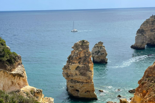 Blick auf das Meer. Man sieht blauen Himmel, Felsen und ein Segelboot.