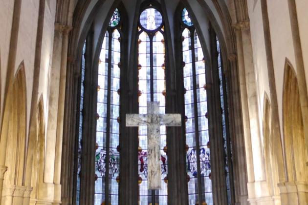 Man sieht auf die gotischen Fenster im Chorraum der Kirche, davor hängt ein Kreuz.