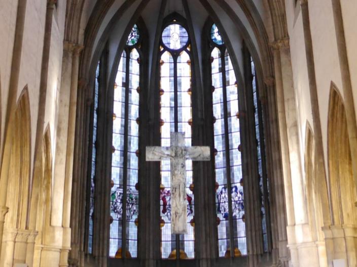 Man sieht auf die gotischen Fenster im Chorraum der Kirche, davor hängt ein Kreuz.