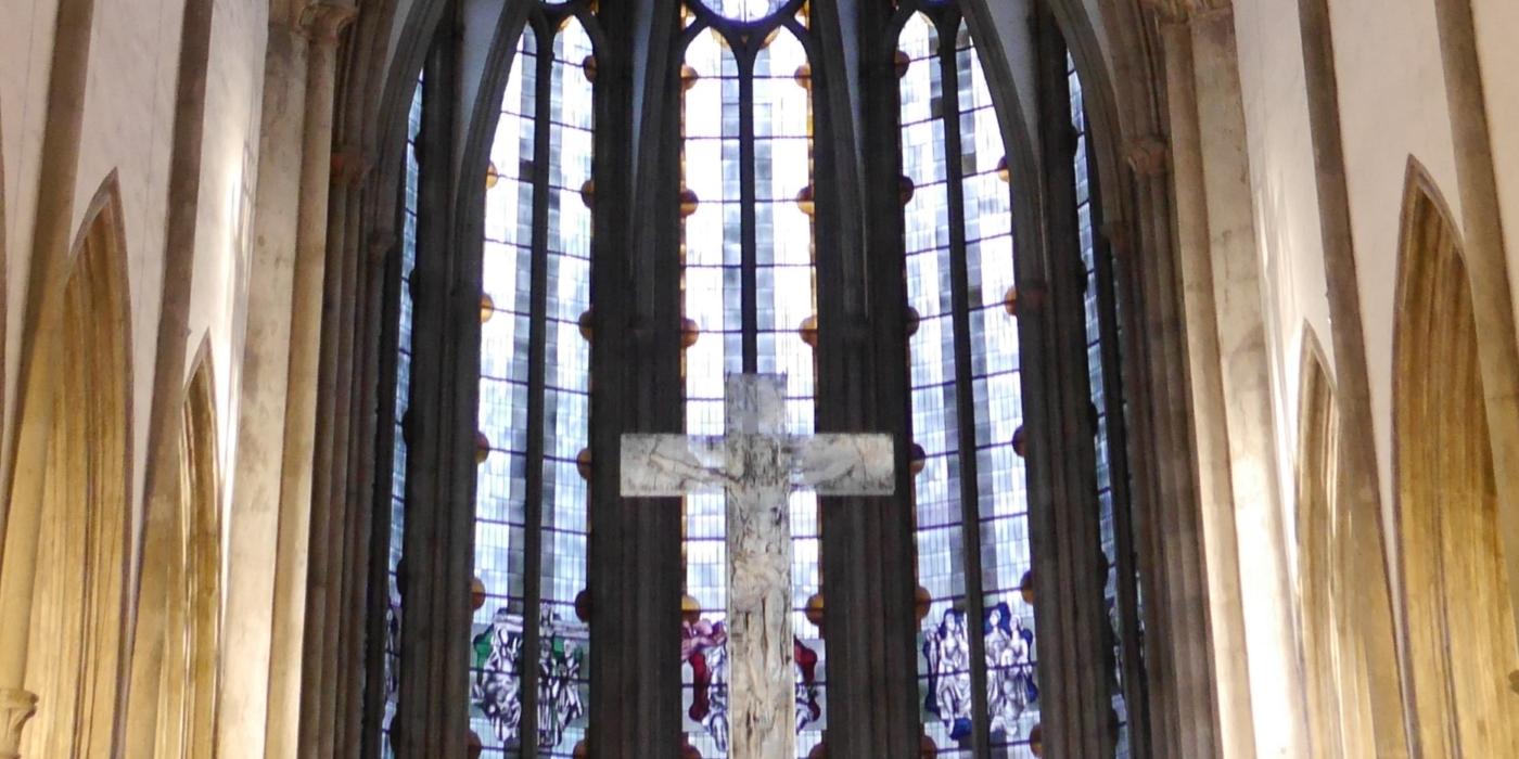 Man sieht auf die gotischen Fenster im Chorraum der Kirche, davor hängt ein Kreuz.
