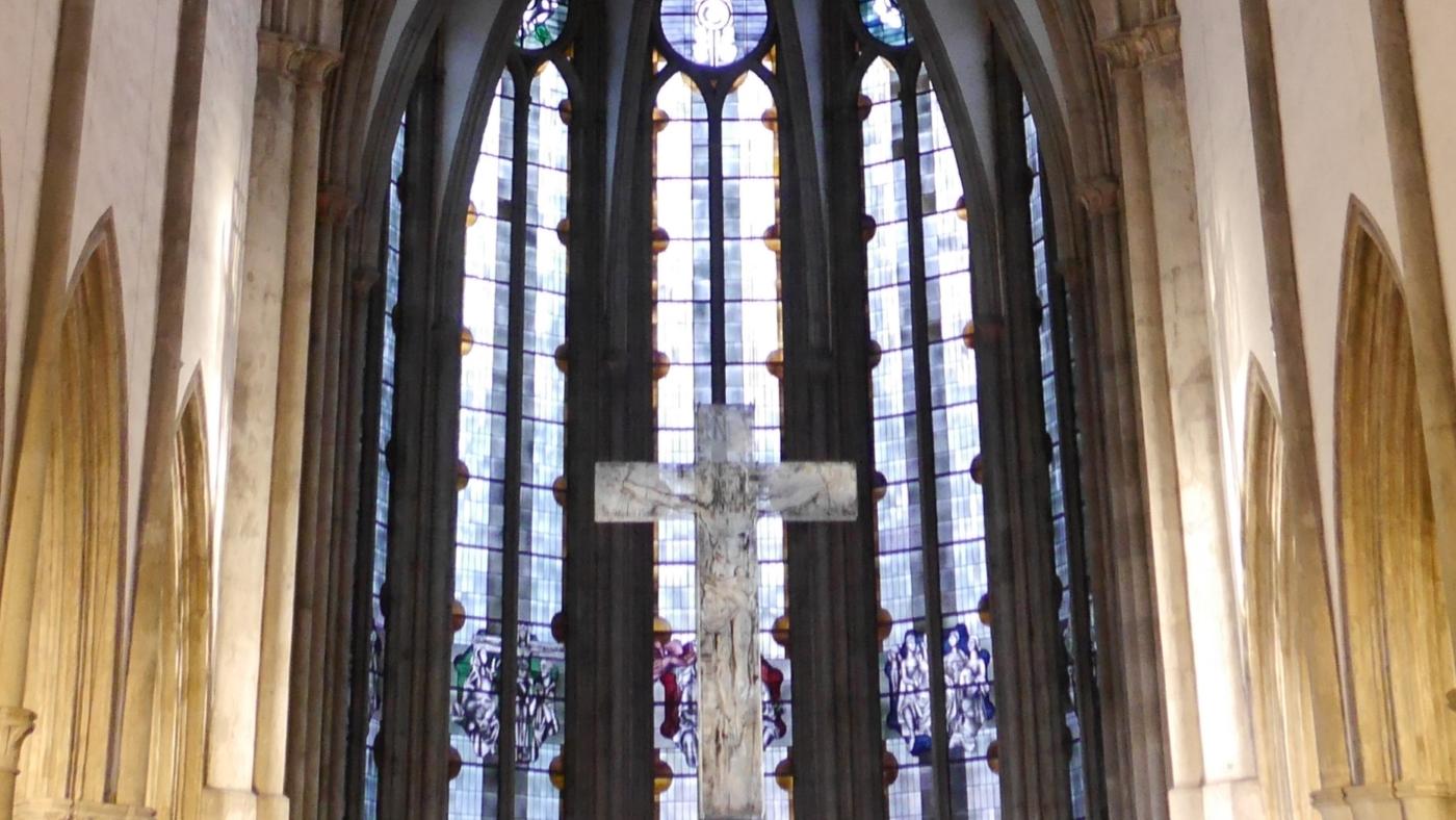 Man sieht auf die gotischen Fenster im Chorraum der Kirche, davor hängt ein Kreuz.