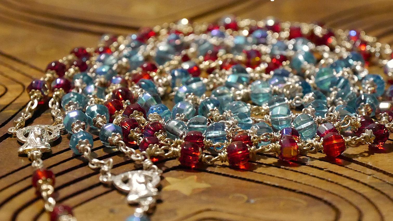 Eine rote und eine blaue Rosenkranz-Kette liegen spiralförmig auf einer Holzunterlage.
