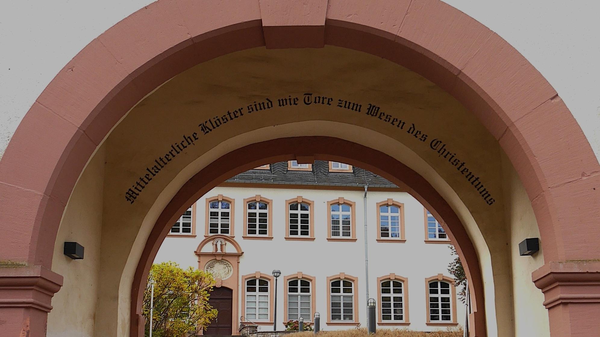 Durch einen Torbogen blickt man auf das Exerzitienhaus, im Torbogen steht 'Mittelalterliche Klöster sind wie Tore zum Wesen des Christentums'.