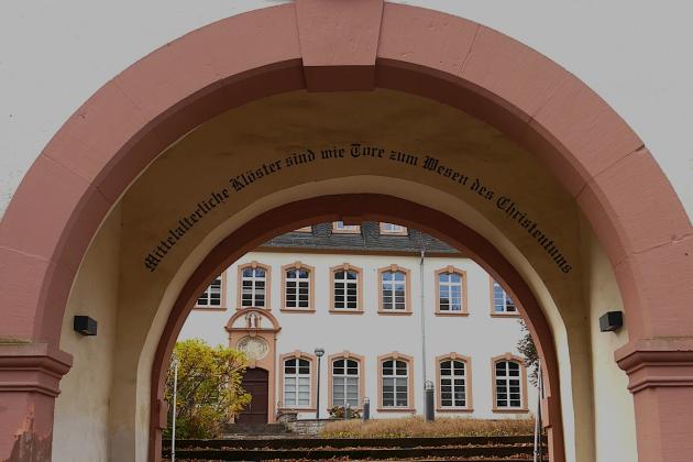 Durch einen Torbogen blickt man auf das Exerzitienhaus, im Torbogen steht 'Mittelalterliche Klöster sind wie Tore zum Wesen des Christentums'.