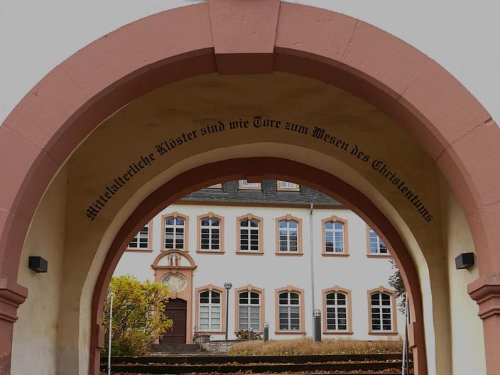 Durch einen Torbogen blickt man auf das Exerzitienhaus, im Torbogen steht 'Mittelalterliche Klöster sind wie Tore zum Wesen des Christentums'.