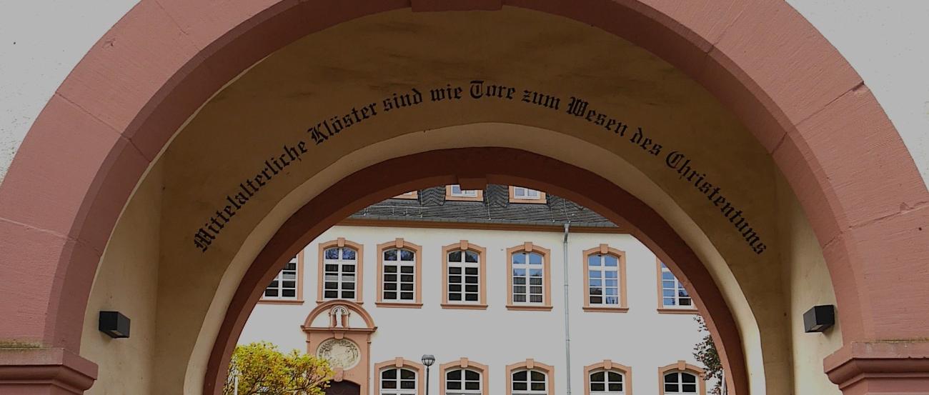 Durch einen Torbogen blickt man auf das Exerzitienhaus, im Torbogen steht 'Mittelalterliche Klöster sind wie Tore zum Wesen des Christentums'.