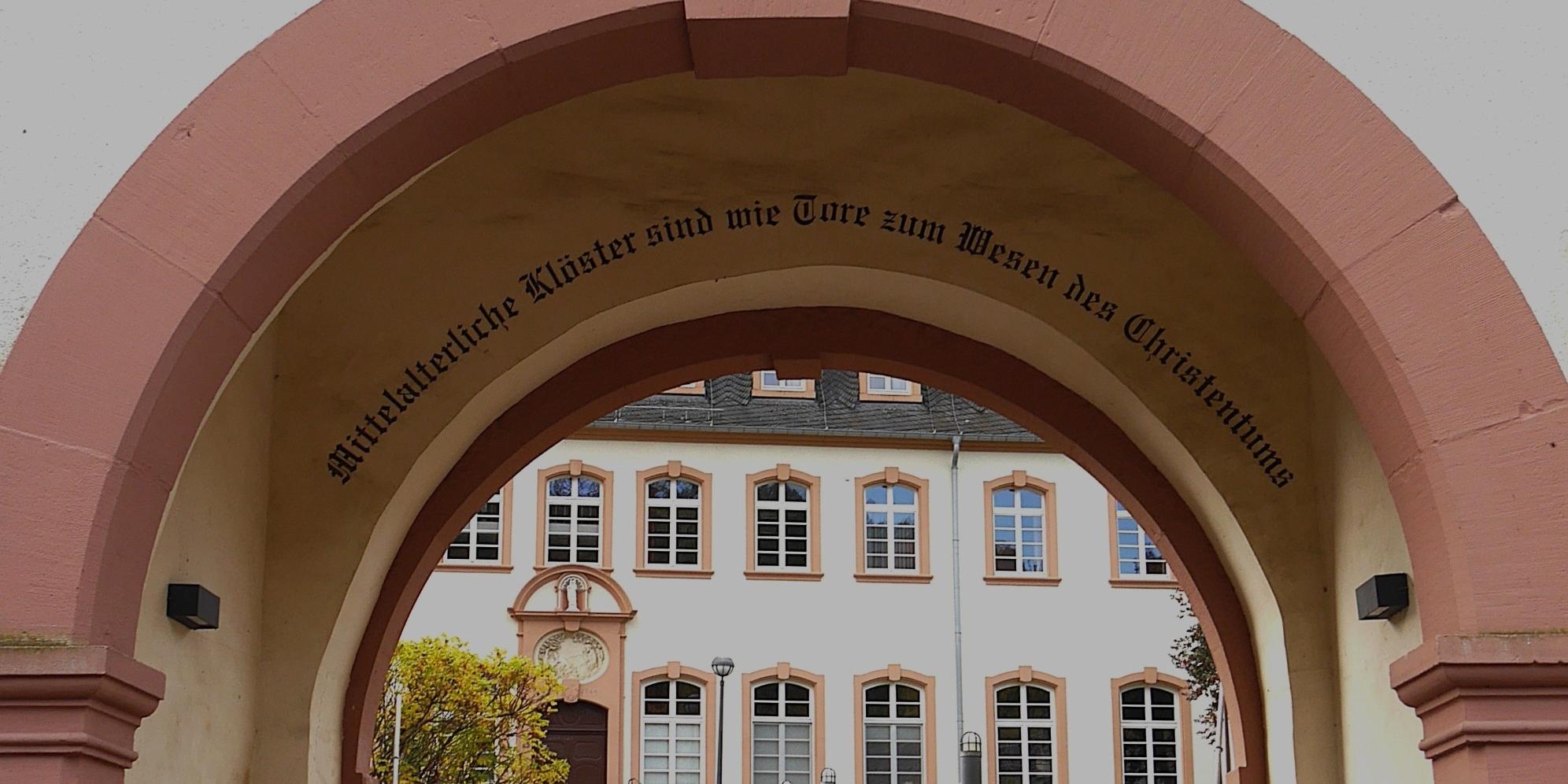 Durch einen Torbogen blickt man auf das Exerzitienhaus, im Torbogen steht 'Mittelalterliche Klöster sind wie Tore zum Wesen des Christentums'.