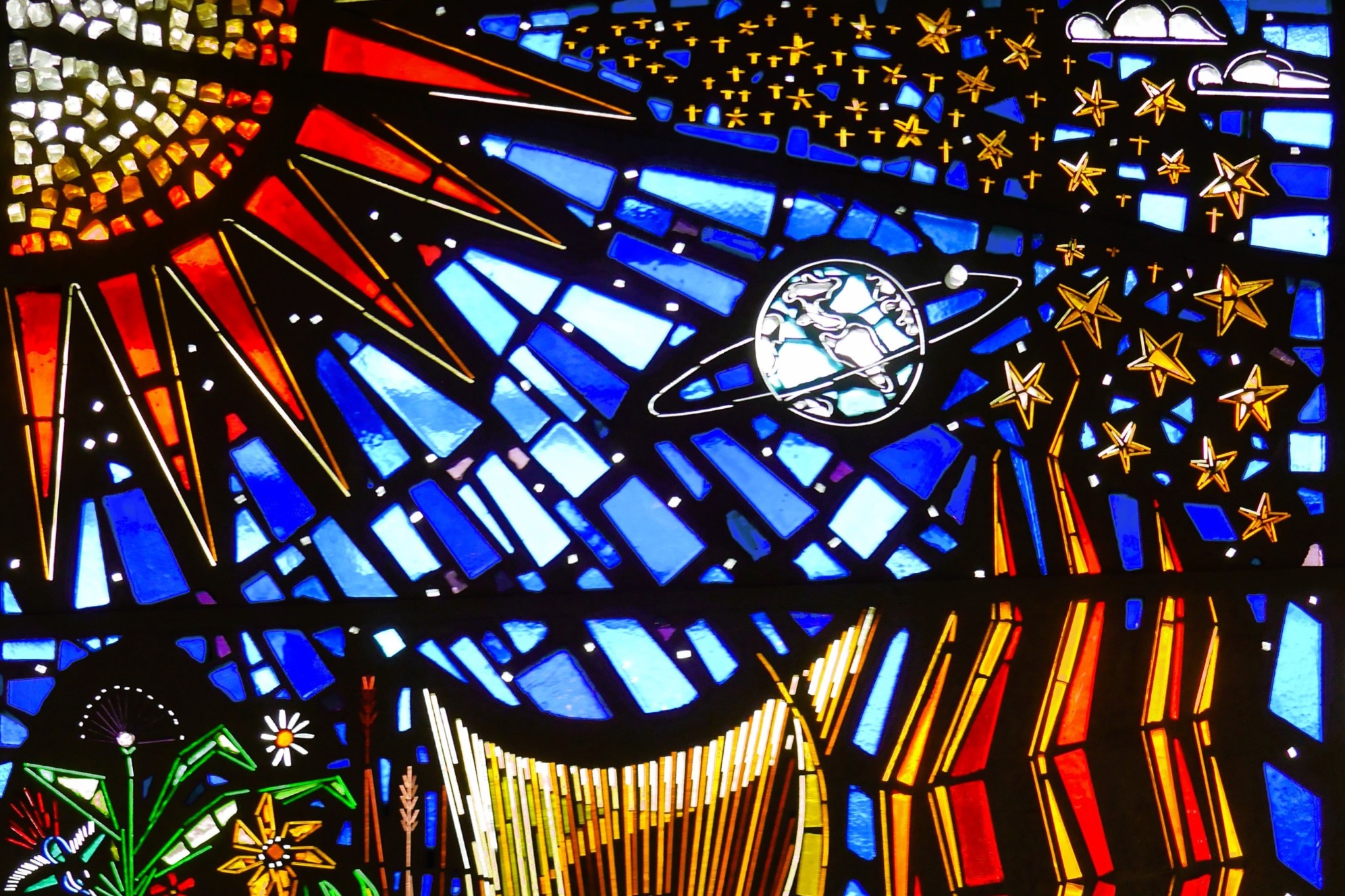 Das Bild zeigt ein Buntglasfenster mit Abbildungen von Sonne, Sternen, Planeten und Pflanzen vor blauem Hintergrund.