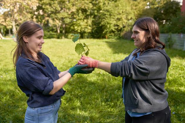 zwei Frauen stehen auf der Wiese, eine Frau gibt der anderen eine kleine Pflanze