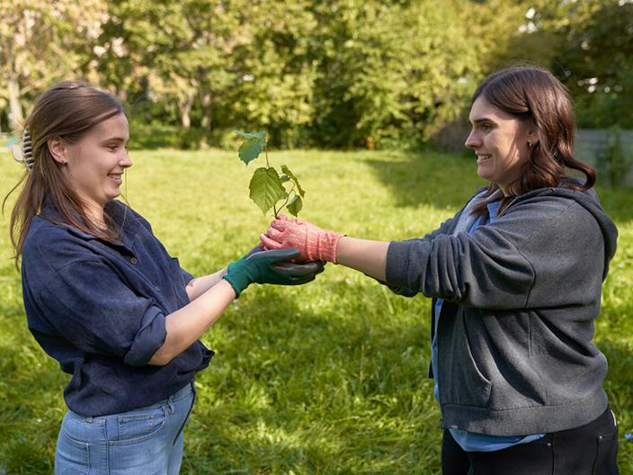 zwei Frauen stehen auf der Wiese, eine Frau gibt der anderen eine kleine Pflanze