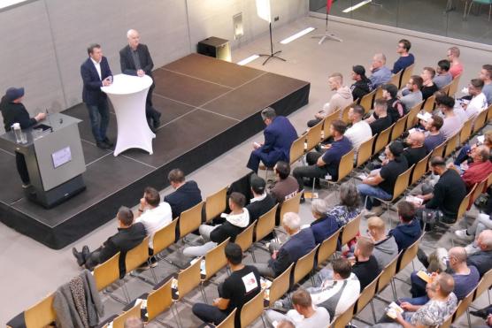 Blick von oben auf die Veranstaltung, man sieht mehrere Reihen mit Menschen, die den Rednern auf dem Podium zuhören.
