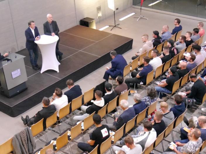 Blick von oben auf die Veranstaltung, man sieht mehrere Reihen mit Menschen, die den Rednern auf dem Podium zuhören.