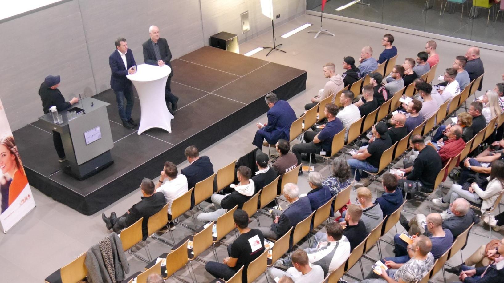 Blick von oben auf die Veranstaltung, man sieht mehrere Reihen mit Menschen, die den Rednern auf dem Podium zuhören.