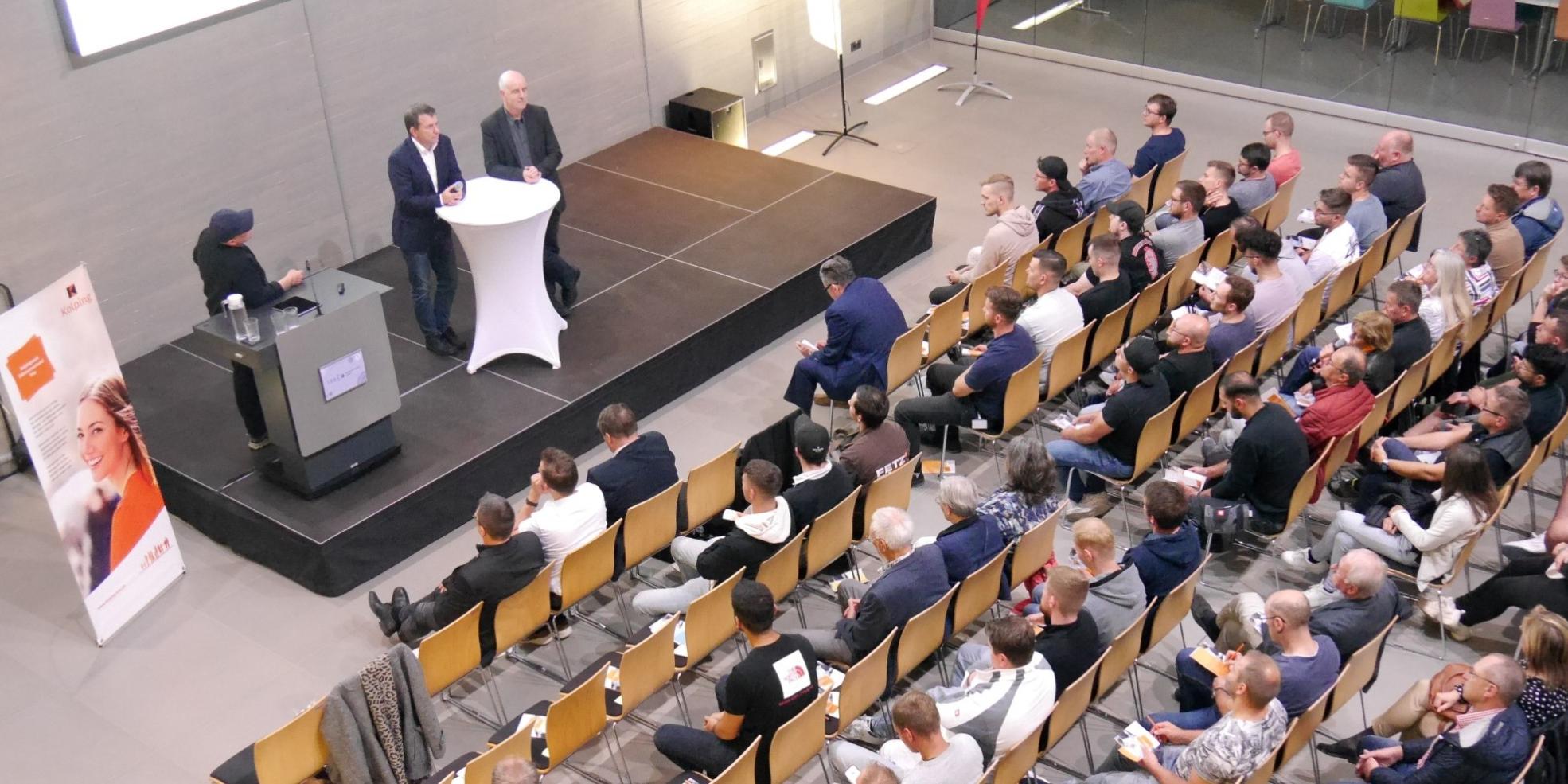 Blick von oben auf die Veranstaltung, man sieht mehrere Reihen mit Menschen, die den Rednern auf dem Podium zuhören.